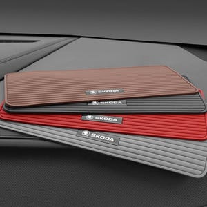 May include: A stack of four rectangular car dashboard mats in brown, black, red, and gray. Each mat features a horizontal ribbed texture and a small, centered rectangular badge with the white text SKODA and the brand logo on a black background.