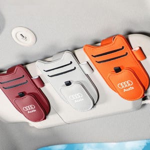 Könnte beinhalten: Drei Auto-Sonnenblenden-Organizer in Kastanienbraun, Hellgrau und Orange, jeweils mit Audi-Logo, zwei horizontalen Kartenschlitzen, einer kleinen Tasche mit Klappe und einer Stiftschlaufe, befestigt an einer hellgrauen Sonnenblende.