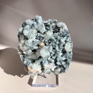 May include: A cluster of pale blue and clear crystal formations, with hints of peach and tan, sits on a clear acrylic stand. The crystals have a rough, textured appearance, with various shapes and sizes. The background is a soft, neutral color.