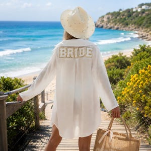 May include: A white long-sleeved button-down shirt with the word BRIDE in gold-trimmed letters on the back, paired with a white straw hat adorned with white pearls. A tan woven tote bag with a ribbon handle is held in the lower right corner.
