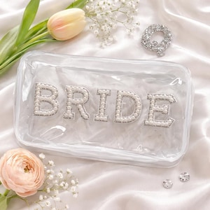 May include: A clear rectangular cosmetic bag with the word "BRIDE" spelled out in pearl and rhinestone letters. The bag is on a white satin surface, with a tulip and other flowers in the background.