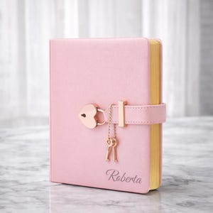 May include: A pink diary with a heart-shaped lock and two small keys hanging from a chain. The diary has gold-colored edges and the name "Roberta" is printed on the cover.