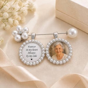 Könnte beinhalten: Eine silberfarbene Brosche mit einer Blume aus weißen Perlen, einem großen Perlenabschluss und zwei runden Anhängern. Ein Anhänger zeigt den Text "Forever in my heart Always by my side" mit einem kleinen Herz, der andere ein Porträt einer Person.