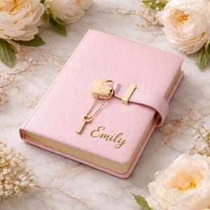 May include: A pink diary with a heart-shaped lock and key. The diary has a gold-colored clasp and the name "Emily" printed in gold. The diary is surrounded by white flowers.