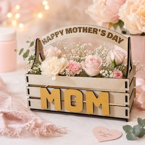 May include: A wooden crate filled with pink and cream-colored flowers, including roses and carnations. The crate has the word "MOM" in gold letters on the front and "HAPPY MOTHER'S DAY" on the top. A heart-shaped card with text is visible.