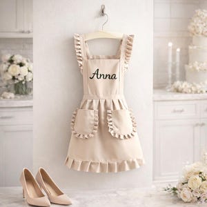 May include: A beige apron with the name "Anna" in black script, hanging on a wooden hanger. The apron has ruffled edges on the straps and pockets. A pair of beige high heels are in the foreground.