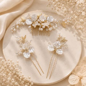 May include: A gold-toned hair comb and two matching hair pins featuring white petal-shaped accents, small clear crystals, and clusters of white pearls arranged on thin, flexible wire branches. The set displays a floral design with metallic leaves.