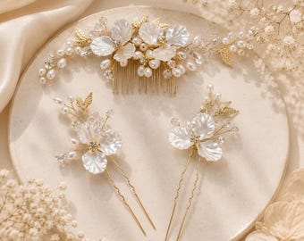 May include: A gold-toned hair comb and two matching hair pins featuring white petal-shaped accents, small clear crystals, and clusters of white pearls arranged on thin, flexible wire branches. The set displays a floral design with metallic leaves.