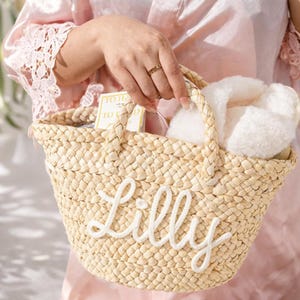 May include: Woven straw tote bag with the name "Lilly" embroidered in white script. The bag has two handles and is filled with white fluffy slippers and other items. The bag is held by a person wearing a pink robe.