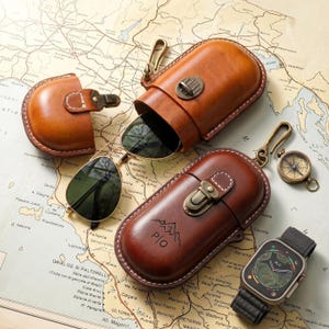 May include: A selection of brown leather accessories, including a sunglasses case, a watch, and a compass, displayed on a vintage map. The sunglasses have gold frames and green lenses. The watch has a dark green strap and face. The case has a mountain logo.