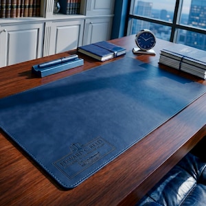 May include: A navy blue leather desk pad with the text "EDWARD C. REED ATTORNEY & COUNSELOR AT LAW" embossed on the lower left corner. A pen, pen holder, clock, and notebooks are on the wooden desk.