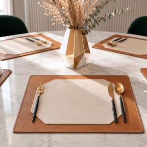 May include: A rectangular placemat with a tan leather-like border and a cream-colored octagonal center. A gold-toned fork, knife, and spoon with black handles are placed on the mat. A geometric gold and white vase sits in the center of the table.