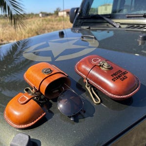 May include: Two brown leather glasses cases with brass-coloured metal hardware and clips. One case is open, revealing a pair of black sunglasses inside. The other closed case features the embossed text PROUD STANFORD DAD on the front surface.