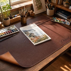 May include: A dark brown leather desk mat with a textured surface and the name David embossed on the right side. The mat features a folded corner revealing a lighter tan underside, providing a workspace for a watercolor painting and brushes.