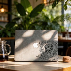 May include: A laptop with a decorative sticker featuring a dog portrait within a floral wreath. The sticker includes the text "Fidèle Ami". The laptop is on a wooden table, with a glass carafe and a macaron in the foreground.