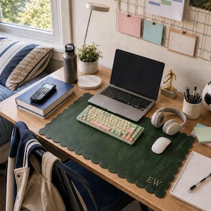 May include: A dark green scalloped desk mat featuring the gold embroidered initials EW in the bottom right corner. The mat is placed on a wooden desk surface, supporting a laptop, a mechanical keyboard with pastel keys, and a white computer mouse.