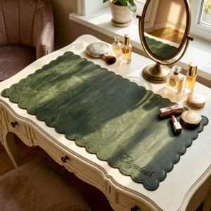 May include: A green leather desk mat with a scalloped edge, personalized with the name "Elizabeth". The mat is placed on a white vanity table, with various beauty products and a mirror.