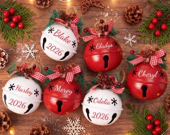 Könnte beinhalten: Sechs runde Weihnachtsbaumkugeln aus Metall in Rot und Weiß, personalisiert mit den Namen Blake, Gladys, Cheryl, Harley, Meroy und Odelia. Einige tragen die Jahreszahl 2026, verziert mit rot-weiß karierten Schleifen, Tannenzapfen und Tannengrün.