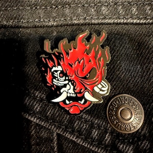 May include: An enamel pin with a red and black oni mask design, complete with flames and white fangs. The pin is set against a dark denim jacket. The design has a skull-like appearance.