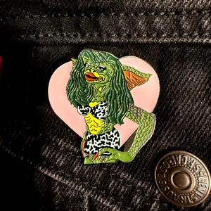 May include: A green and yellow enamel pin featuring a female Gremlin character with green hair, a leopard print bikini, and a pink heart background. The pin is set against a black denim jacket.
