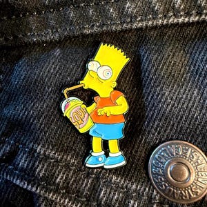 May include: A vibrant enamel pin of Bart Simpson enjoying a milkshake. Bart is shown in his iconic yellow skin, orange top, and blue shorts. The milkshake cup has the word "Squishee". The pin is set against a dark denim background.