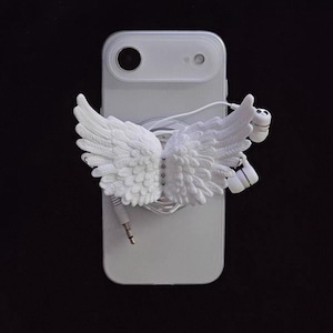 Op de afbeelding: Een witte telefoonhoes met een ontwerp van gebeeldhouwde engelen vleugels. De vleugels zijn bevestigd aan de achterkant van de telefoonhoes. Witte bedrade oortelefoons zijn aangesloten op de telefoon. De achtergrond is zwart.