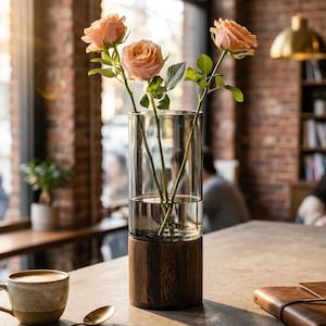 May include: A cylindrical glass vase with a dark brown wooden base containing three peach-colored roses with green stems and leaves. The vase is filled with water and sits on a flat surface next to a ceramic coffee mug and a metal spoon.
