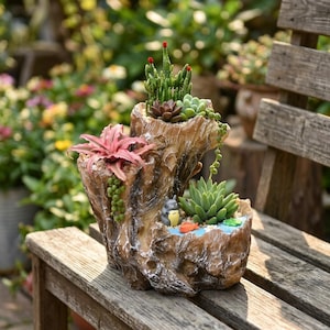 Op de afbeelding: Een meerlaagse bruine hars plantenbak in de vorm van een boomstronk, voorzien van een roze bromelia, groene vetplanten, een klein grijs Totoro-figuurtje en een piepklein oranje visje als decoratief accent.