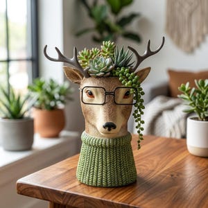 May include: A deer-shaped planter featuring a brown textured head with dark antlers, wearing black-rimmed glasses and a green knit-patterned base. The top of the head holds a variety of green succulents, including a trailing string of pearls plant.