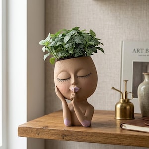 May include: A tan-colored head-shaped planter featuring a face with closed eyes, long eyelashes, and a finger held to the lips in a shushing gesture. The lips are painted light purple, and the planter holds a green plant with white-veined leaves.
