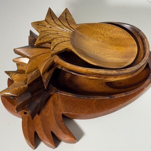 May include: A set of wooden serving bowls and spoons, carved in the shape of pineapples. The bowls and spoons are made of a warm brown wood and are stacked together. The pineapple design includes detailed leaf and crown carvings.