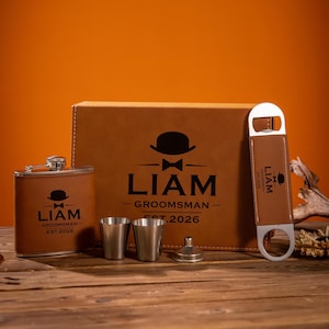 May include: A brown leatherette groomsmen gift set with a flask, two shot glasses, a funnel, and a bottle opener. The set is personalized with the name "LIAM" and the text "GROOMSMAN EST. 2026" in black.