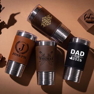 May include: Four stainless steel insulated tumblers with personalised leather sleeves. One is brown with "ETHAN" in gold, another brown with "THOMAS" and crossed swords, a third with "JOSEPH" and a laurel wreath, and a black one with "DAD EST 2026".