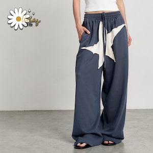 May include: Dark blue wide-leg sweatpants with a large white star design on the front. The pants have a drawstring waist and pockets. The pants are made of a soft material.