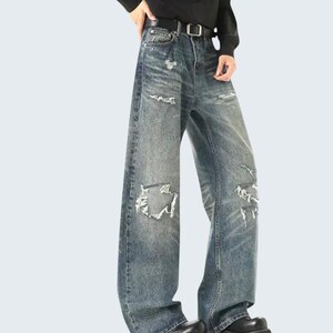May include: Faded blue denim jeans with distressed details, featuring ripped sections and a wide-leg design. The jeans have a classic five-pocket style and are worn with a black belt.