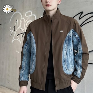 Könnte beinhalten: Braune Jacke mit blauen Jeans-Einsätzen an den Ärmeln. Die Jacke hat einen hohen Kragen, einen Frontreißverschluss und einen kleinen rechteckigen Aufnäher auf der Brust. Der Hintergrund ist eine graue Wand mit Graffiti.