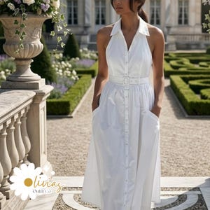 Puede incluir: Un vestido blanco con cuello halter, botones delanteros, cinturón y bolsillos. El vestido llega a la rodilla y tiene cuello. El vestido se usa al aire libre en un jardín. El vestido es de Olivia's Outfit Co.
