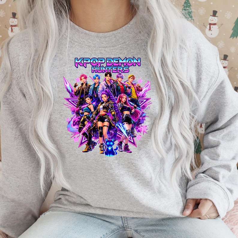 May include: A light gray sweatshirt featuring a vibrant graphic of a group of anime-style characters. The graphic includes the text "KPOP DEMON HUNTERS" in a neon-style font. The characters are depicted in various poses with colorful outfits.