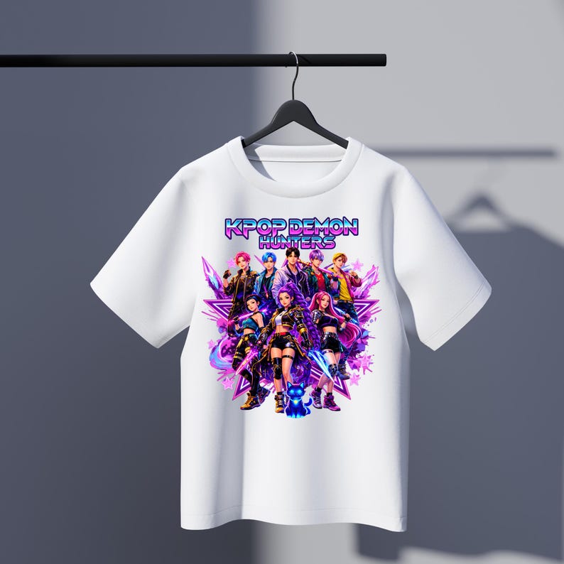 May include: White t-shirt featuring a graphic of a group of anime-style characters with the text "KPOP DEMON HUNTERS" in a vibrant, colorful design. The design includes stars and a dynamic, action-oriented composition.