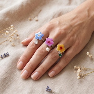 Könnte beinhalten: Drei silberne Ringe mit baumelnden Blumenanhängern. Der erste Ring hat eine blaue Blume mit einem klaren Kristall, der zweite eine rosa Blume mit einer weißen Perle und der dritte eine gelbe Blume mit einem grünen Edelstein. Jeder Ring hat eine dünne Schiene.