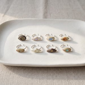 May include: Eight silver rings with open bands, each featuring a unique, small, smooth, pebble-like stone or ceramic accent. The stones vary in color and pattern, including shades of white, brown, and grey, with some displaying small painted dots or lines.