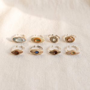 May include: Eight adjustable metal rings featuring unique, small, hand-formed ceramic focal pieces. Each ceramic element displays distinct shapes, textures, and color accents, including shades of blue, brown, white, and tan, arranged in two rows.