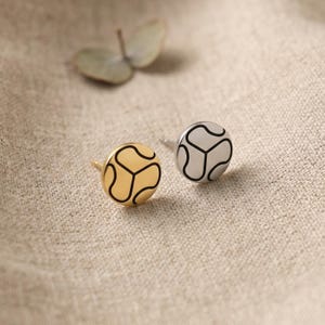 May include: A pair of stud earrings featuring a geometric pattern. One earring is gold-coloured with a black outline, the other is silver-coloured with a black outline. The earrings are circular and rest on a beige fabric.