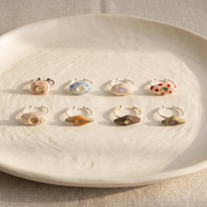 May include: Eight adjustable metal rings featuring unique ceramic focal points. Each ring displays a distinct design, including speckled textures, blue and white patterns, solid earth tones, and red polka dots, all centred on a thin silver-toned band.