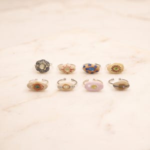 May include: Eight adjustable silver-tone rings, each featuring a unique, small, oval-shaped ceramic or stone cabochon. The cabochons display various hand-painted patterns, including dots, lines, and floral motifs in colors like blue, red, and tan.