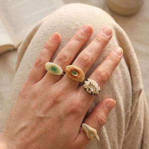 May include: Four rings with distinct ceramic-style focal points worn on fingers. One ring features a pale green center, another has concentric orange and brown rings, a third is white with black speckles, and the fourth is cream with small dots.