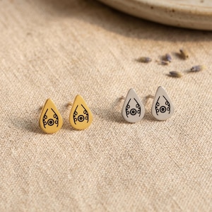 May include: Two pairs of teardrop-shaped stud earrings. One pair is gold-colored, the other is silver-colored. Each earring features a black, stylized design with a central circle and curved lines. The earrings are displayed on a neutral-toned fabric.