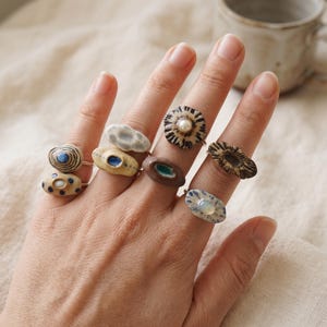 May include: A collection of eight ceramic rings worn on fingers, featuring various organic shapes, textures, and colors including beige, brown, blue, and white. Several rings are accented with small embedded pearls or iridescent stones.