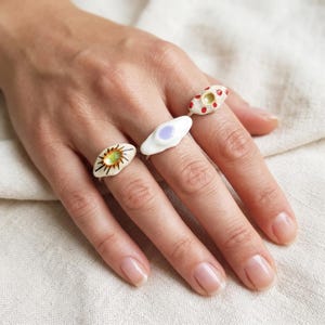 May include: Three rings worn on fingers. The first ring features a cream-colored oval with a central green iridescent eye and black radiating lines. The second is a white oval with a central light purple dot. The third is a cream oval with red dots.