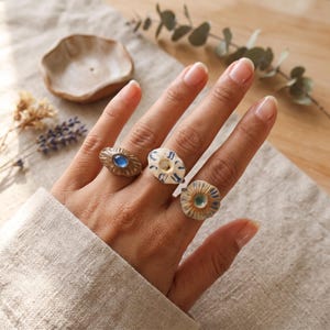 May include: Three rings worn on fingers. The first ring is textured bronze with a central blue oval stone. The second ring is white ceramic with blue painted details. The third ring is a textured, flower-shaped ceramic piece with a blue center.